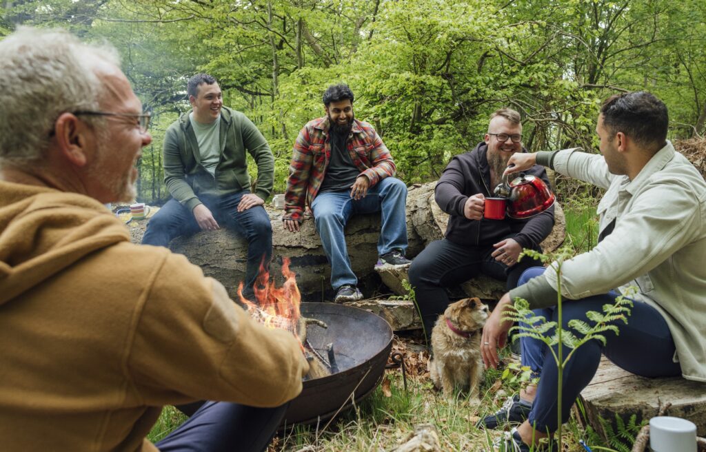 5 men sitting on large logs in the woods around a campfire. One man is pouring coffee into another man's cup.