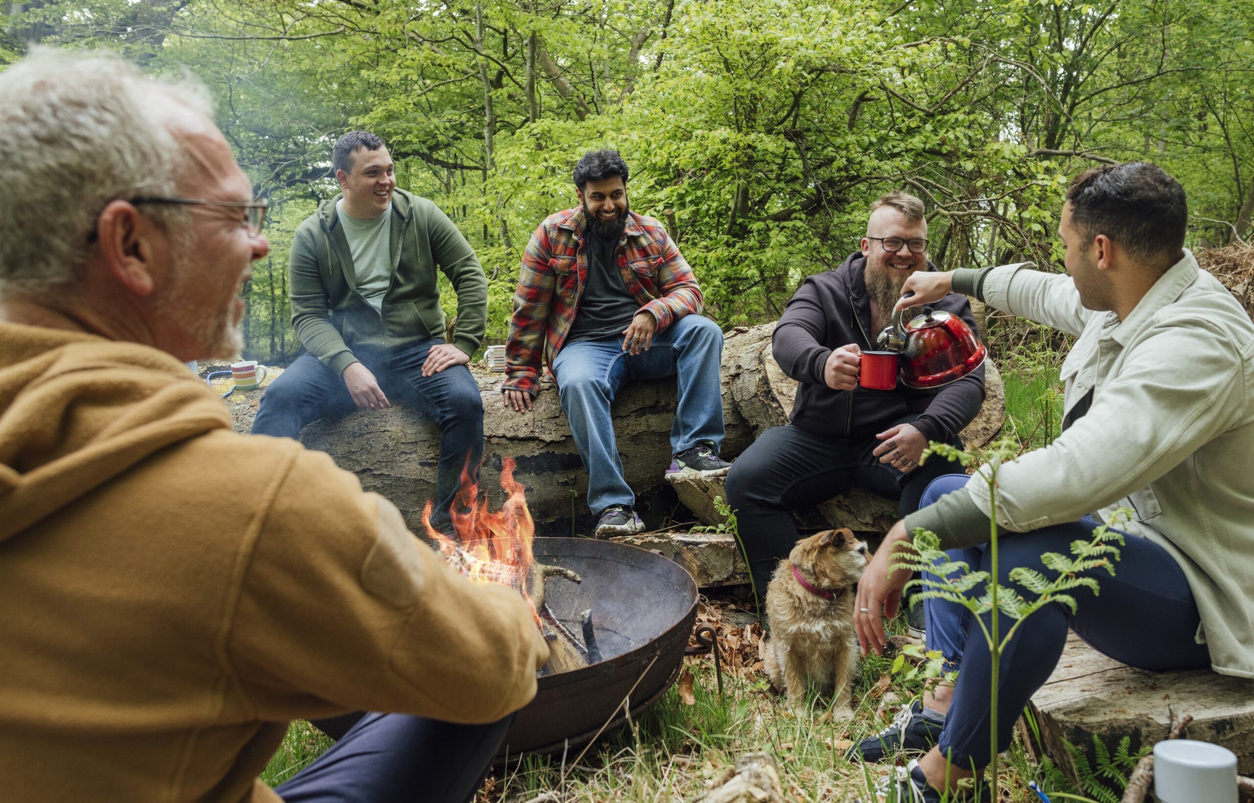 5 men sitting on large logs in the woods around a campfire. One man is pouring coffee into another man's cup.