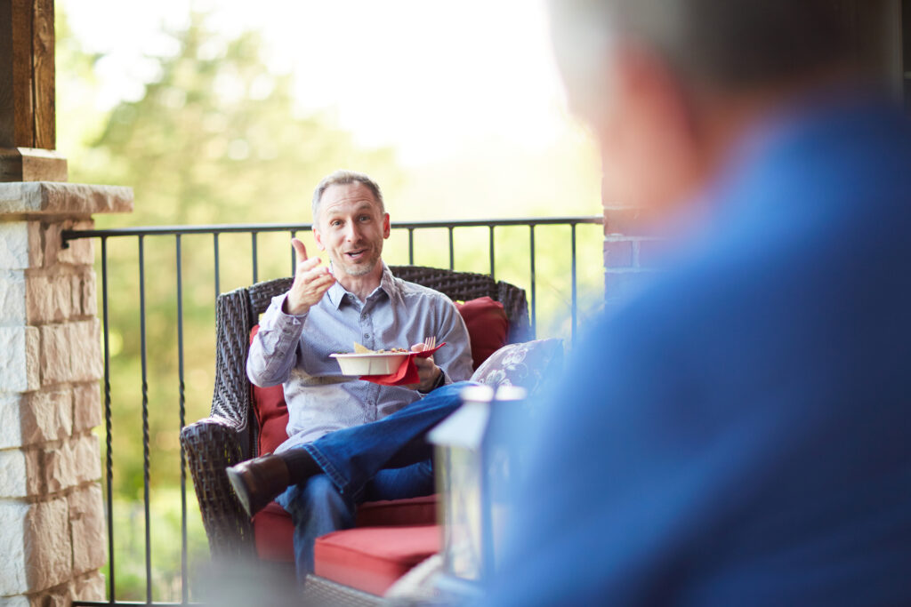 Man sitting on outdoor patio gesturing with one hand and holding a bowl in the other with a blurred man in the foreground.