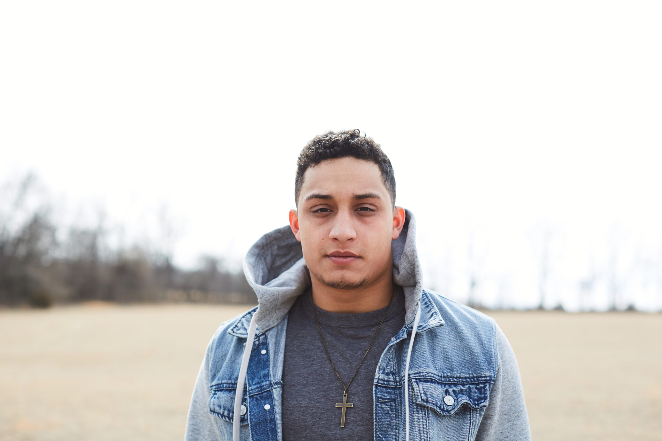 Young man wearing a jean jacket with gray sweatshirt sleeves and hood. He has a cross necklace on over a heather navy shirt.