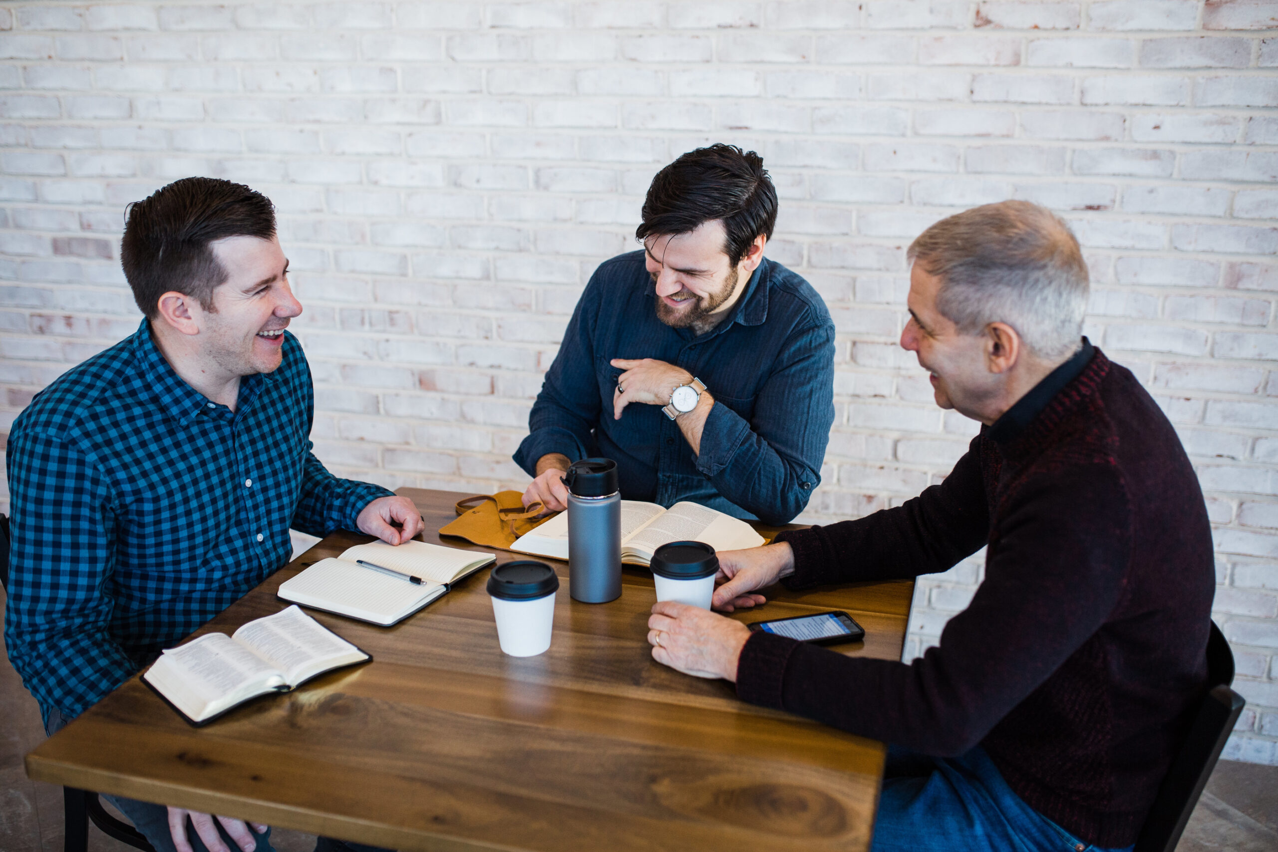 Three men laughing together sitting at a square, wooden table with coffee cups and open bibles on top