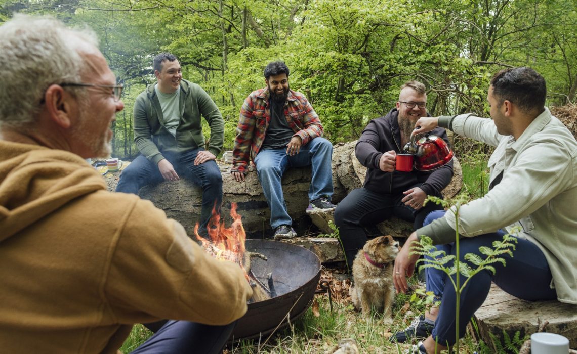 5 men sitting on large logs in the woods around a campfire. One man is pouring coffee into another man's cup.