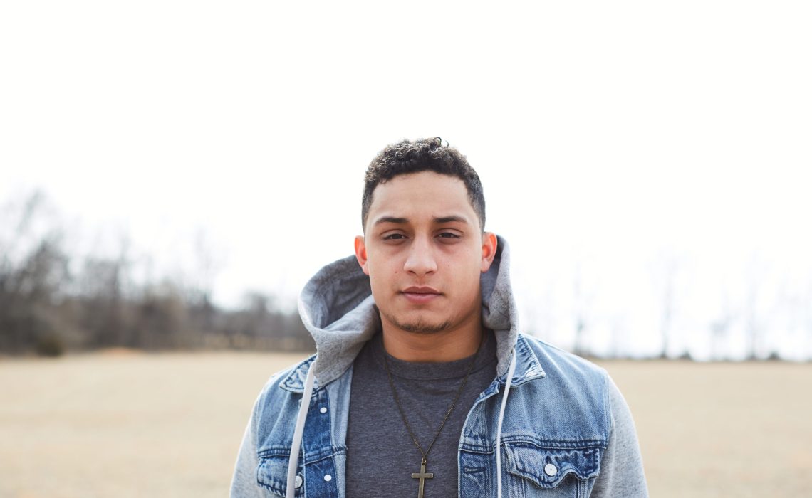 Young man wearing a jean jacket with gray sweatshirt sleeves and hood. He has a cross necklace on over a heather navy shirt.