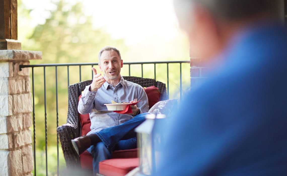 Man sitting on outdoor patio gesturing with one hand and holding a bowl in the other with a blurred man in the foreground.