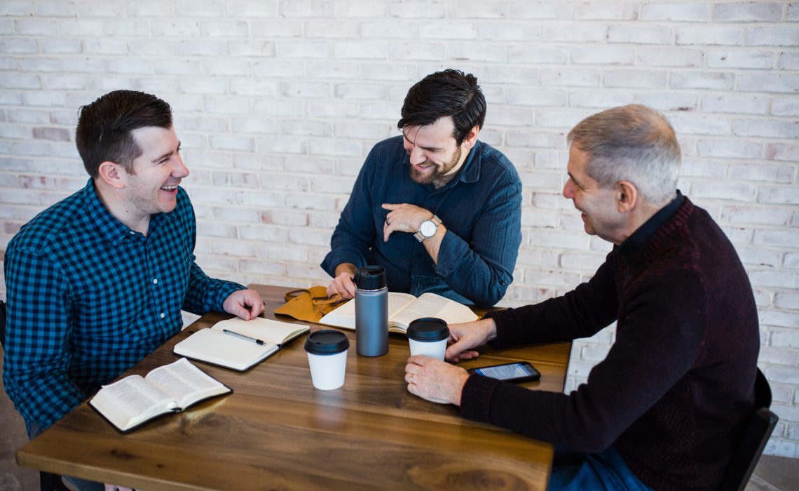 Three men laughing together sitting at a square, wooden table with coffee cups and open bibles on top